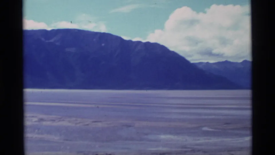 1977 Alaska 8mm frame showing wide flat terrain, distant mountains, and a large white cloud against a pale sky.