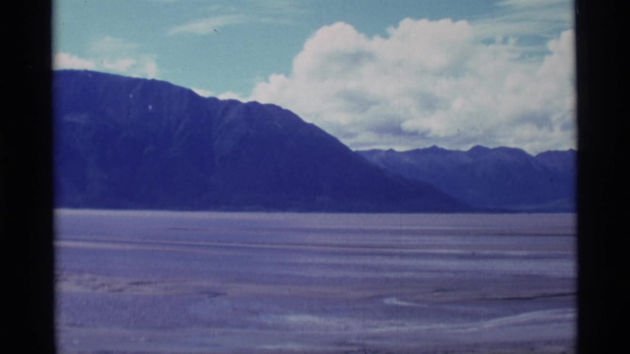 1977 8mm Alaska frame showing dark mountain slopes, a wide flat plain, faint water channels, and a cloudy sky.