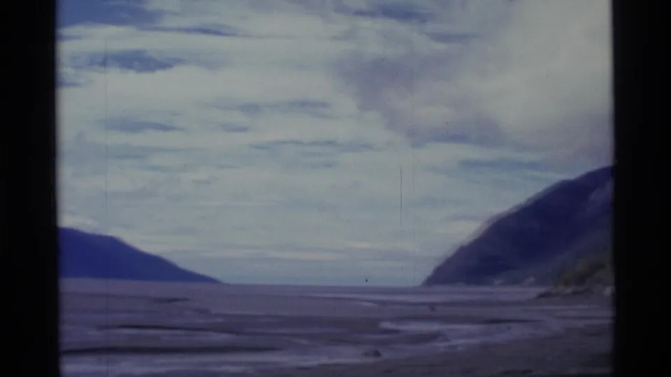1977 8mm Alaska frame showing open flat terrain, faint water channels, low distant landforms, and a dark mountain slope at right.