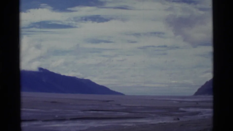 1977 8mm Alaska landscape with distant mountain headlands, a broad flat foreground, muted water traces, and a cloudy sky.