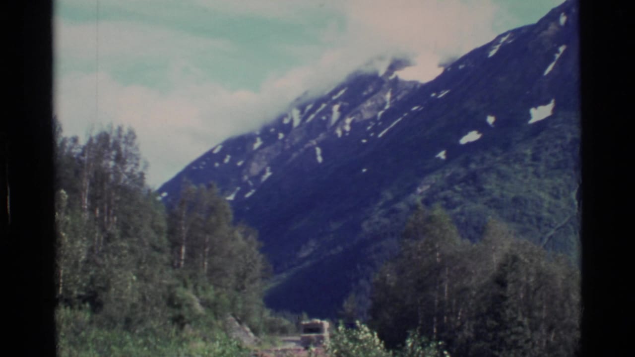 1977 Alaska 8mm frame showing a snow-patched mountain ridge rising behind trees and valley vegetation.