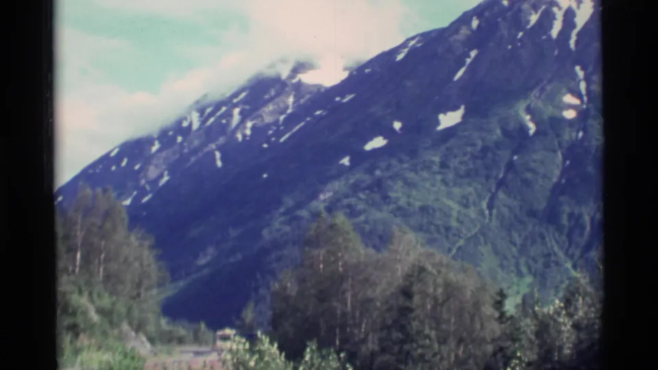 1977 Alaska 8mm frame with a broad mountain slope, scattered snow patches, trees, and soft pale sky.