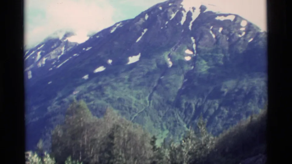 1977 Alaska 8mm frame showing a closer mountain face with green slopes, snow remnants, and treetops below.