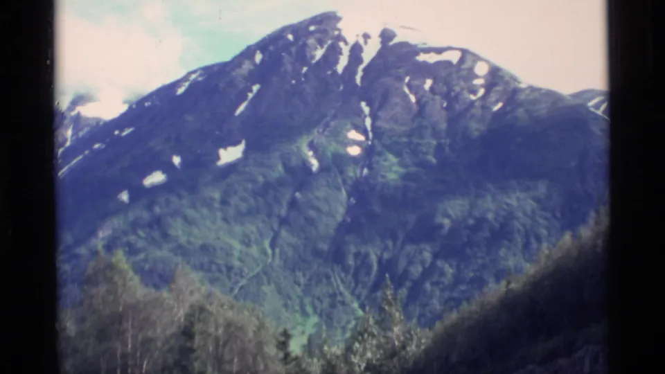 1977 Alaska 8mm frame of a snow-marked mountain peak above forested lower slopes and valley trees.