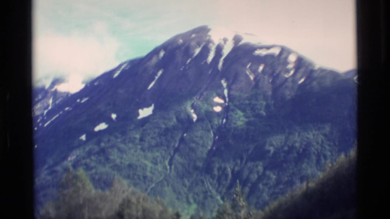1977 Alaska 8mm frame showing a snow-streaked mountain ridge rising above forested lower slopes beneath a pale sky.
