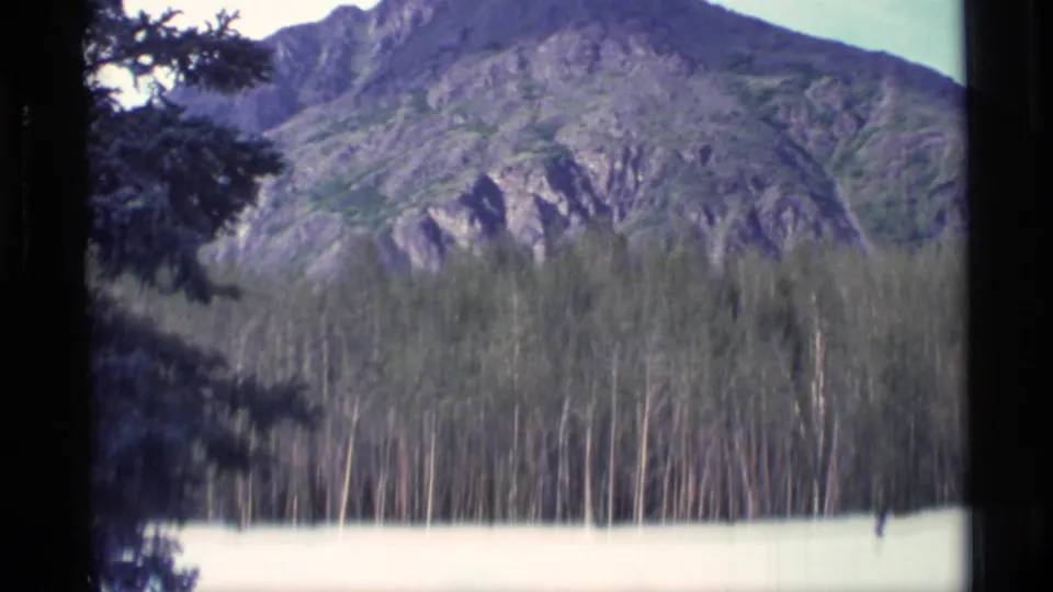 1977 Alaska 8mm frame with rocky mountain slopes, dense trees, and a light-toned lower edge that may be water, snow, or exposure flare.