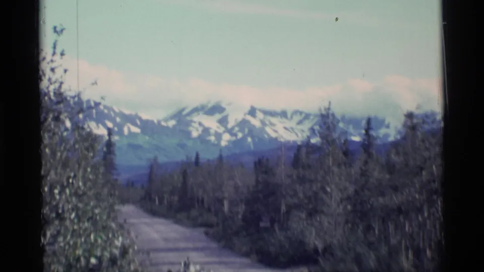 Alaskan home-movie view of a tree-lined road with blue-green mountains and patches of snow beyond the forest.