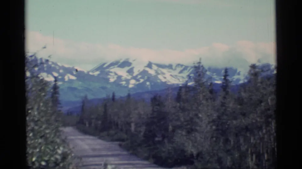 1977 Alaska 8mm landscape frame with roadside trees in the foreground and snow-streaked mountains on the horizon.
