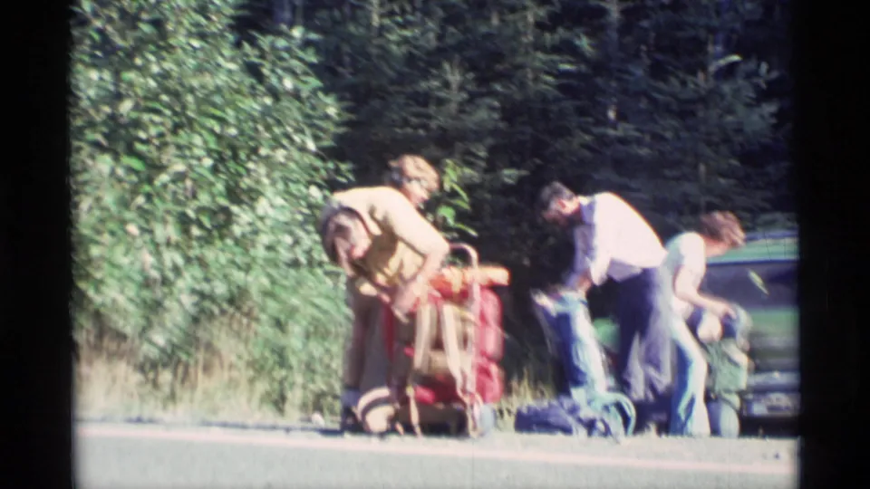 1977 Alaska home-movie frame of three people preparing outdoor gear beside a green vehicle at a trailhead.