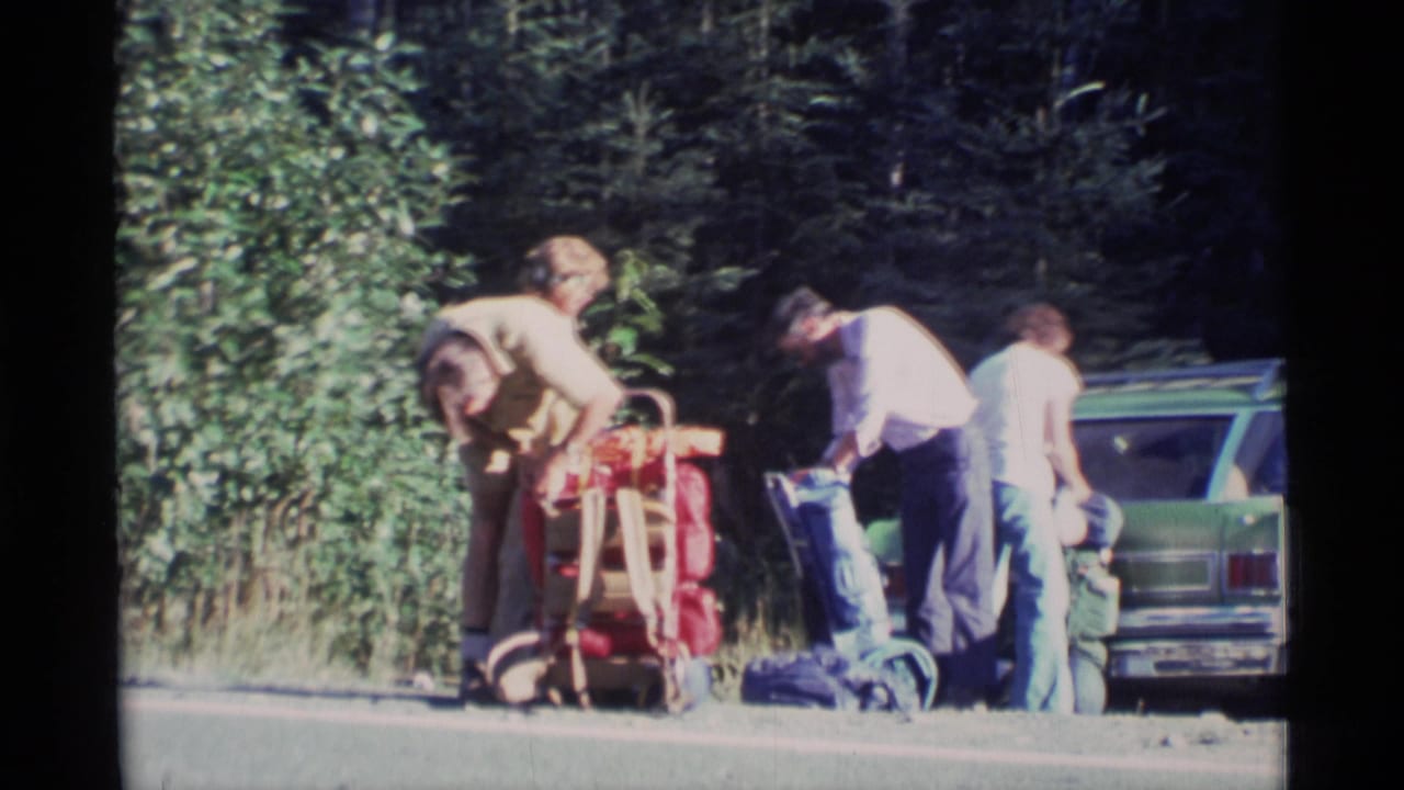 1977 Alaska home-movie frame showing backpackers organizing red and blue packs beside a green station wagon at a wooded roadside pullout.