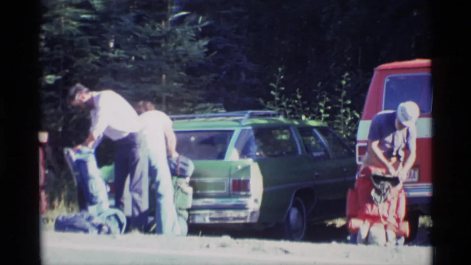 Backpackers in casual 1970s clothing adjust gear between a green station wagon and a red van in an Alaska forest parking area.