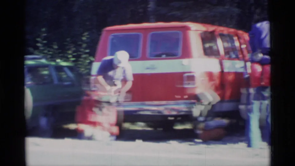 A backpacker bends over a red pack beside a red van, with another loaded pack partly visible in the wooded parking area.