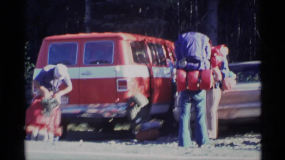 Backpackers prepare large packs beside a red van in an Alaska forest pullout, with gear on the ground and trees behind them.