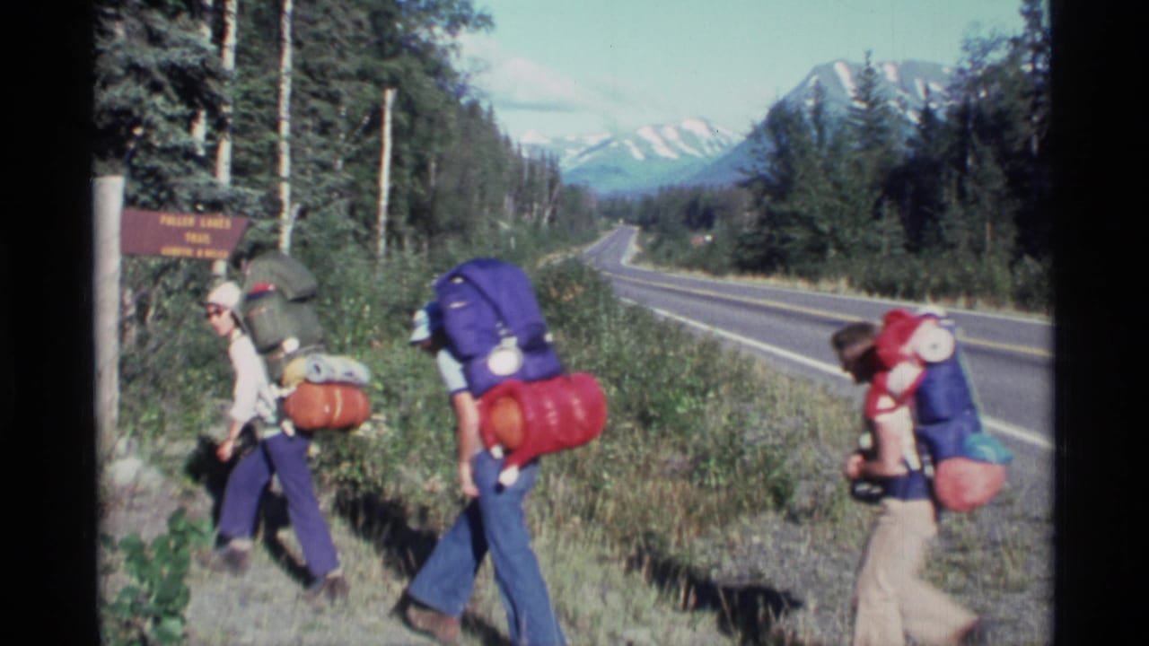 Three backpackers walk past a Fuller Lakes Trail sign beside a road with mountains in the distance.