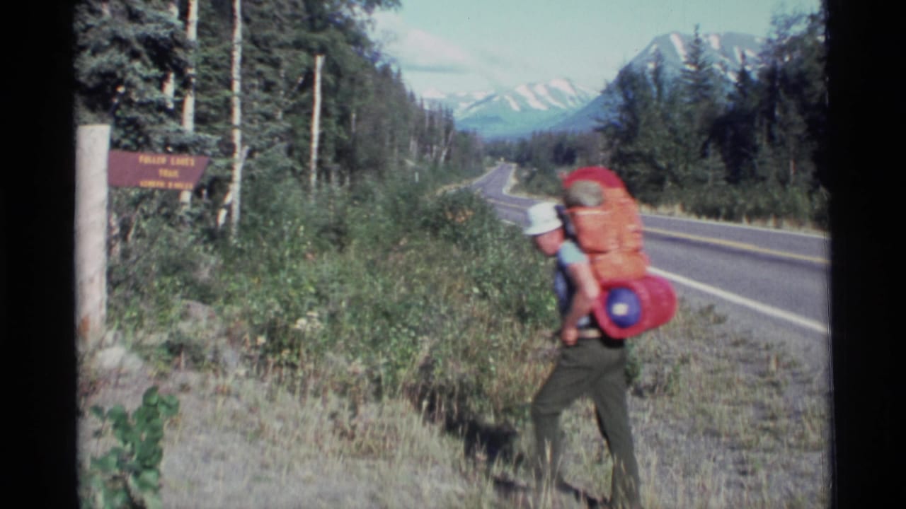 A backpacker in a white shirt carries a red pack along the roadside near the trail sign.