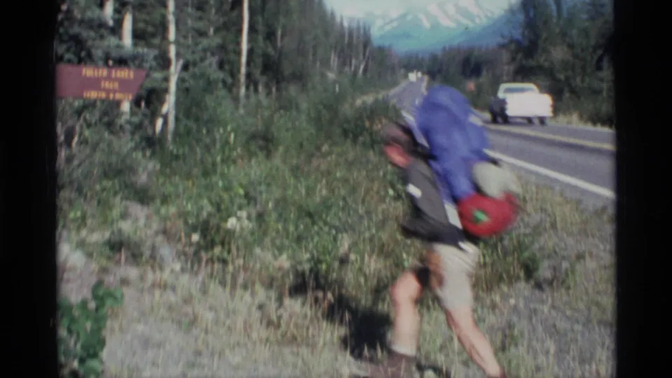 A hiker carrying a blue pack passes through roadside vegetation in Alaska, with a white vehicle and another car on the road behind.