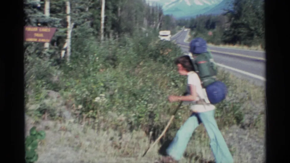 A hiker in light pants carries a large blue backpack and walking stick beside the Alaska roadside, with the Fuller Lakes Trail sign and road visible.
