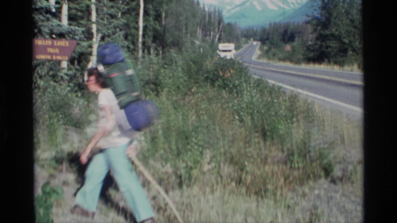 Backpacker walks past a Fulmer Lakes Trail sign beside a paved mountain road.