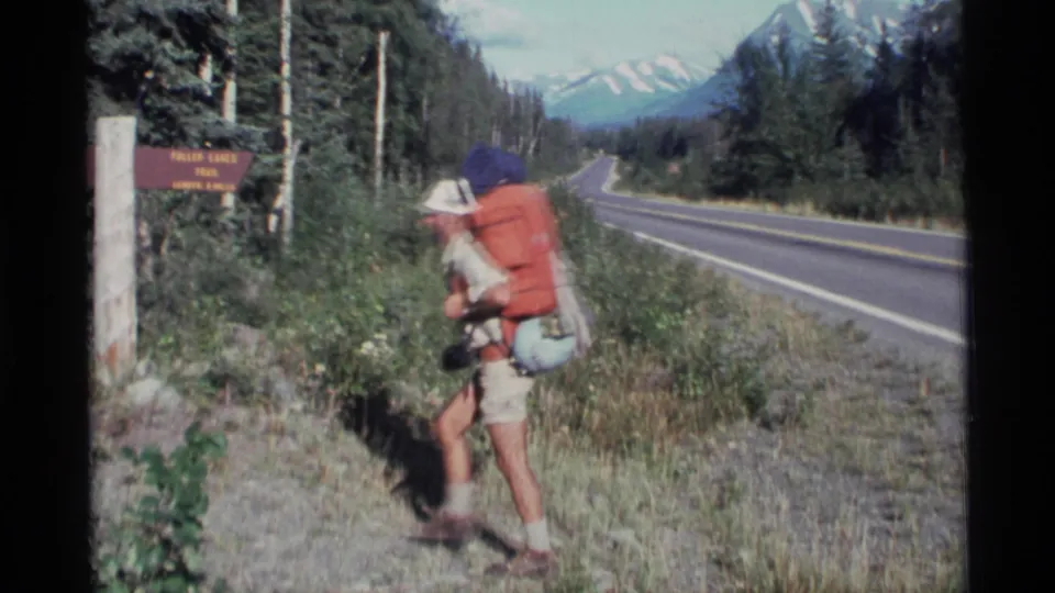 A hiker in a brimmed hat and orange pack moves by the Fulmer Lakes Trail sign in archival Alaska road footage.
