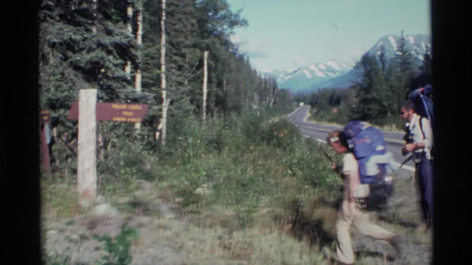 Two backpackers move past the Fulmer Lakes Trail sign near a paved Alaskan road with mountains in the distance.