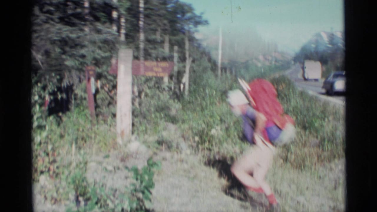 Backpacker bends near a roadside trail sign with highway and mountains in the background.