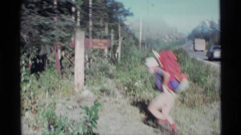 In 1977 Alaska home-movie footage, a backpacker bends near a trail sign beside a highway with forest and distant mountains behind.