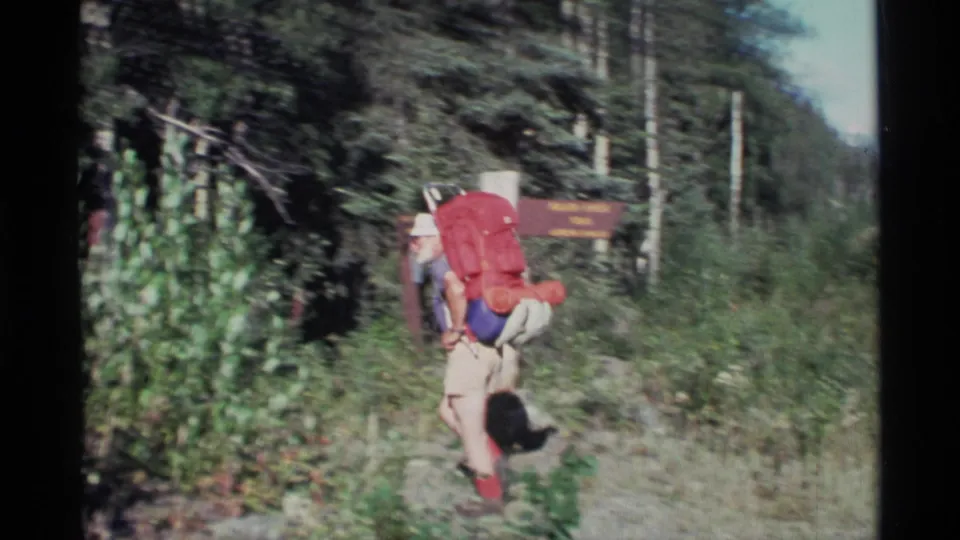 A hiker carrying a large red backpack walks past a trail sign into dense Alaska roadside forest in 1977 footage.
