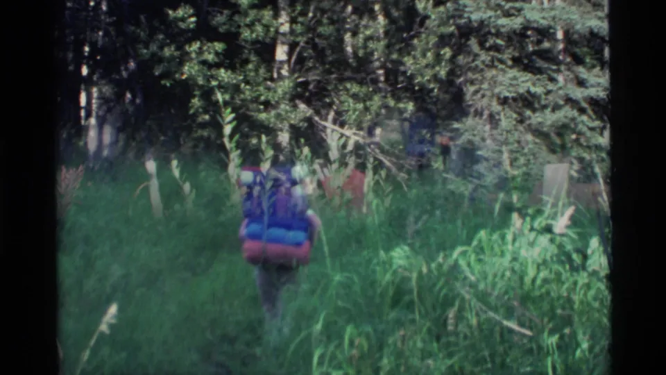 A backpacker seen from behind follows a narrow grassy path into Alaska forest, filmed in handheld 8mm style.