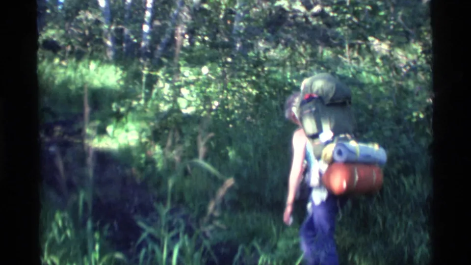 A backpacker in 1977 Alaska walks away through dense forest, carrying a large pack with rolled gear attached.