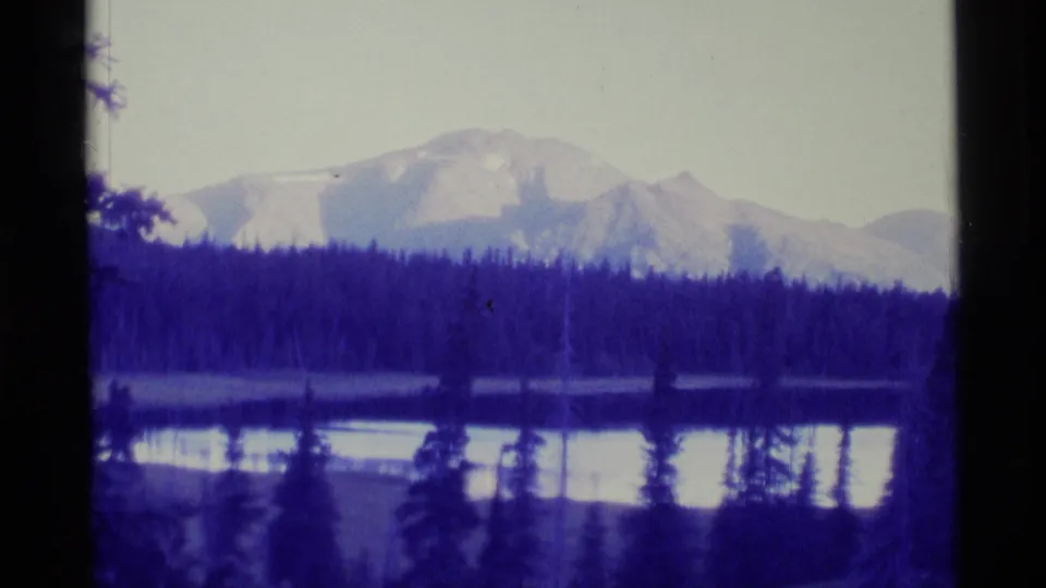 1977 Alaska 8mm frame with a still lake, dark evergreen shoreline, and pale mountain range under a bright sky.