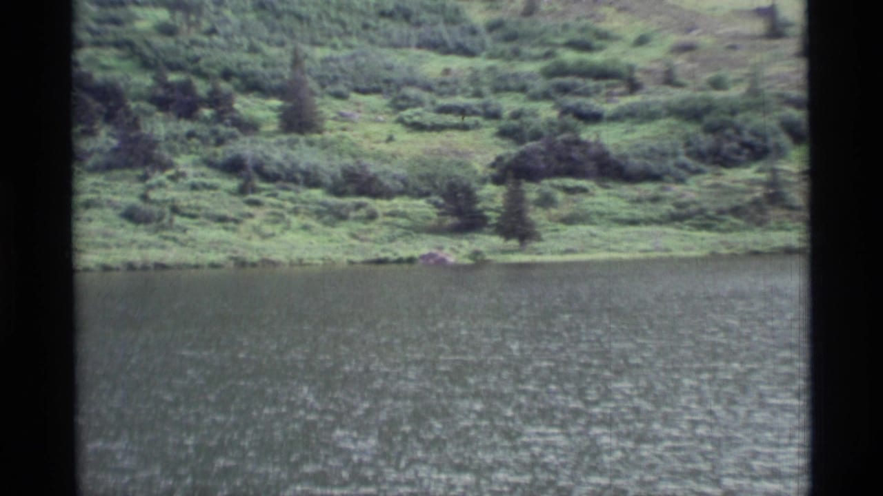 1977 Alaska 8mm frame showing rippled lake water, a green shoreline, shrubs, and scattered evergreen trees.