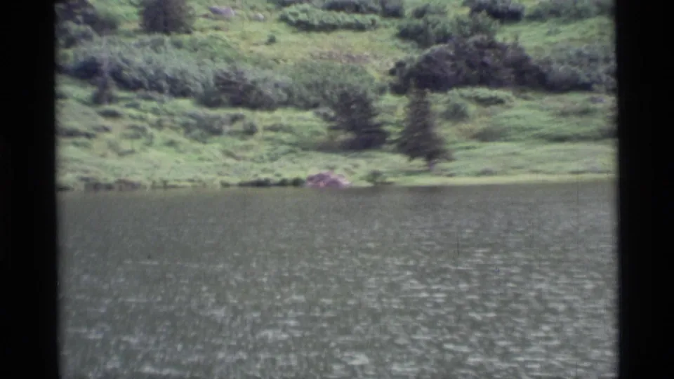 1977 Alaska home-movie frame of a rippled lake bordered by green banks, shrubs, and dark evergreen trees.