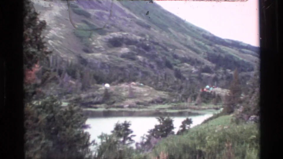 A 1977 Alaska mountain lake view shows evergreen trees, grassy shoreline, and small distant camp-like shapes near the water.