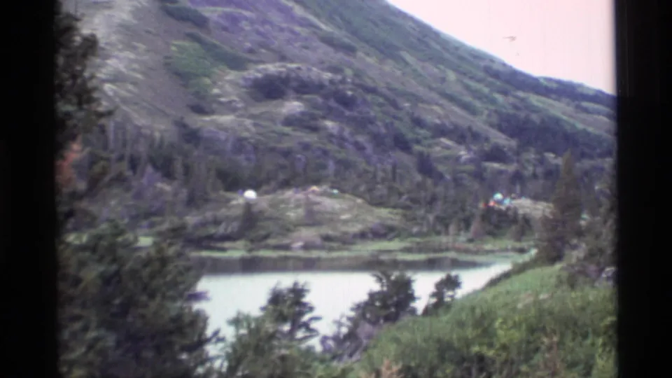 An Alaskan mountain lake appears in soft 8mm color, with wooded shore, grass, and small distant figures or tents that need review.