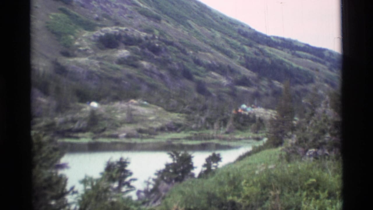 1977 Alaska 8mm frame showing a quiet mountain lake, forested slopes, and a small distant campsite on a grassy rise.