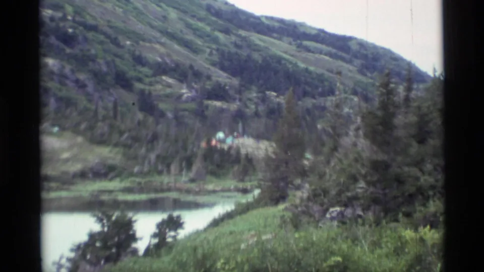 Alaska wilderness view with lake water, dense evergreens, green slopes, and a distant cluster of colorful tents.