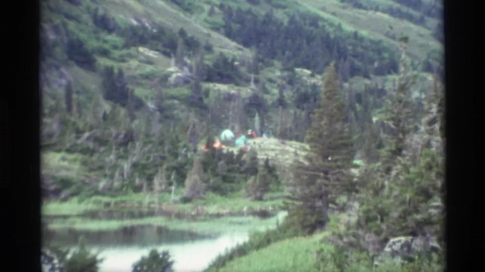 Closer 1977 Alaska 8mm view of colorful tents on a grassy hillside above a quiet lake and evergreen trees.