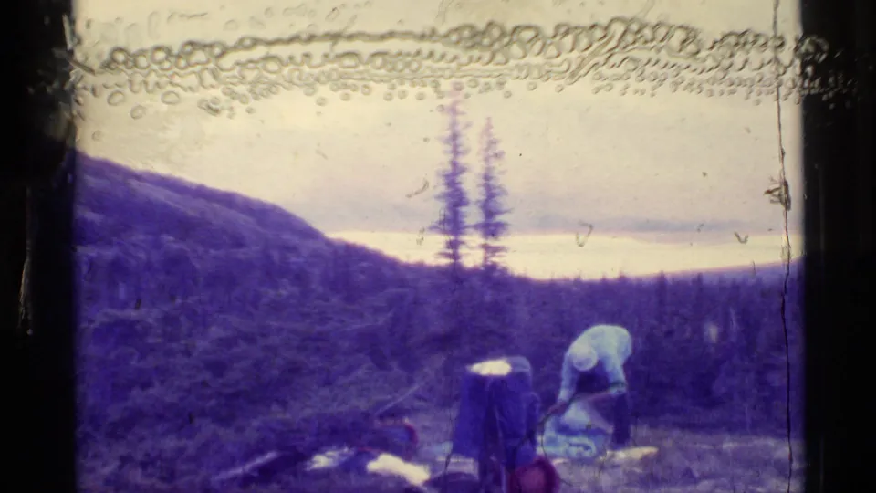 Adult man in a light shirt bends over camping gear on an Alaska hillside, with lake, trees, and hazy mountains beyond.