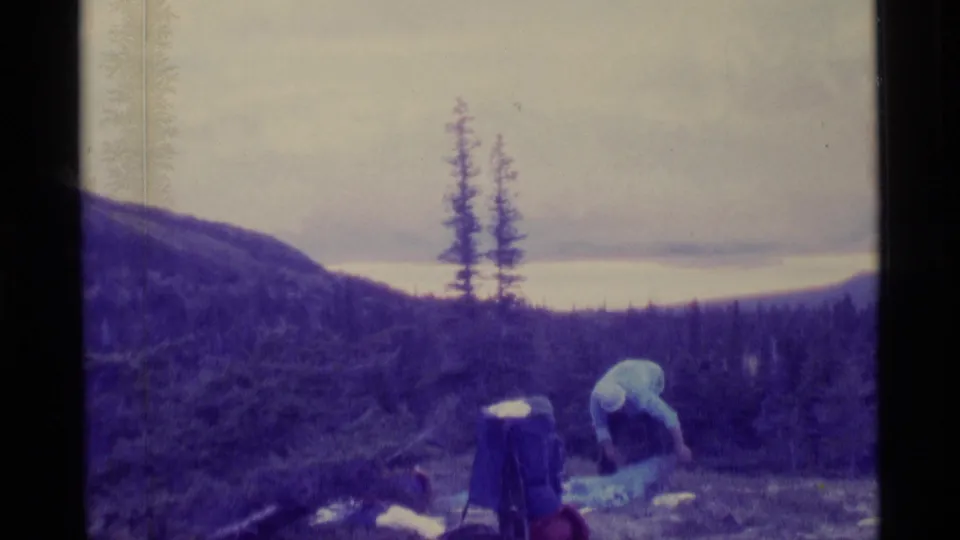 1977 Alaska home-movie frame with an adult leaning over outdoor supplies in a purple-tinted evergreen landscape.