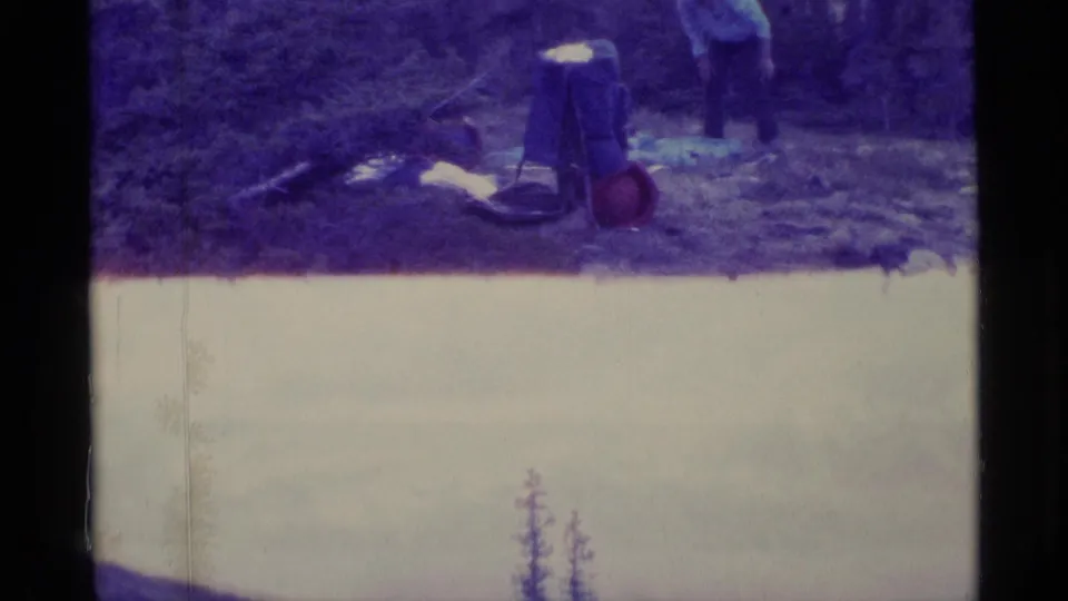 Faded 8mm frame from Alaska showing picnic gear, a bag, and a partly visible adult at the edge of a forest clearing.