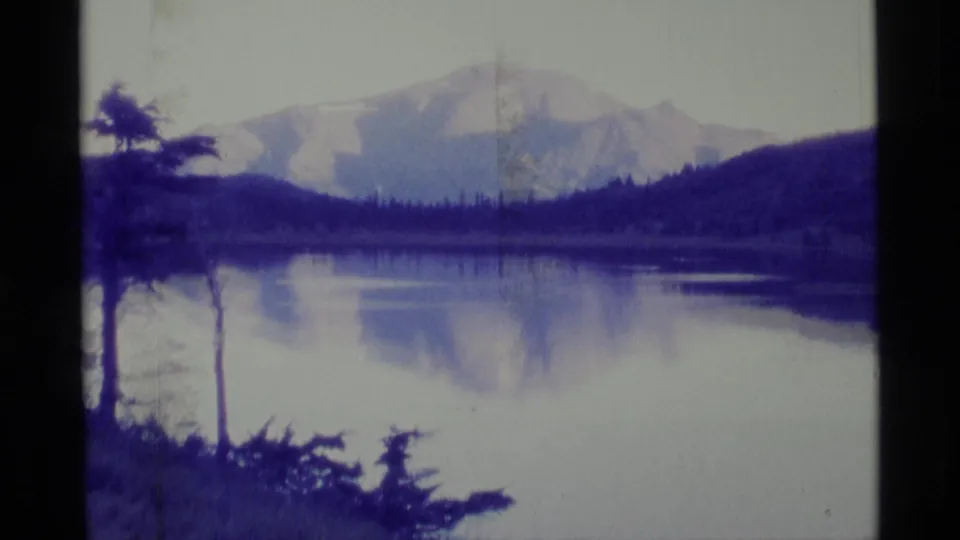 1977 Alaska 8mm frame with a snow-capped mountain and dark hills reflected in calm pond water, with trees at the shore.