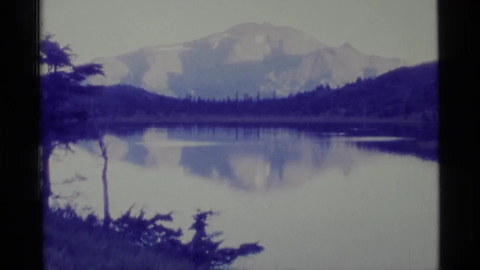 1977 Alaska 8mm view of a mountain reflected across a pond, with evergreen silhouettes and low hills around the water.