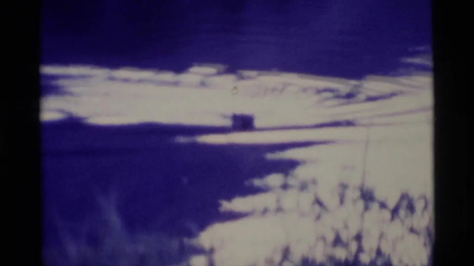1977 Alaska 8mm frame showing reflective pond water with bright sky reflection, ripples, and dark shoreline vegetation.