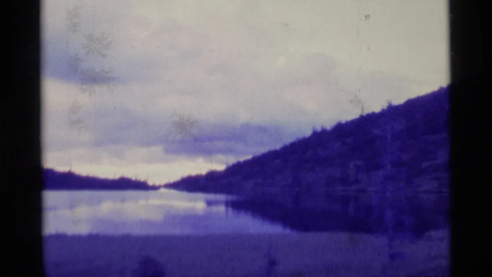 Wide 1977 Alaska 8mm view of water between wooded hills beneath cloudy sky, with visible grain and color fading.