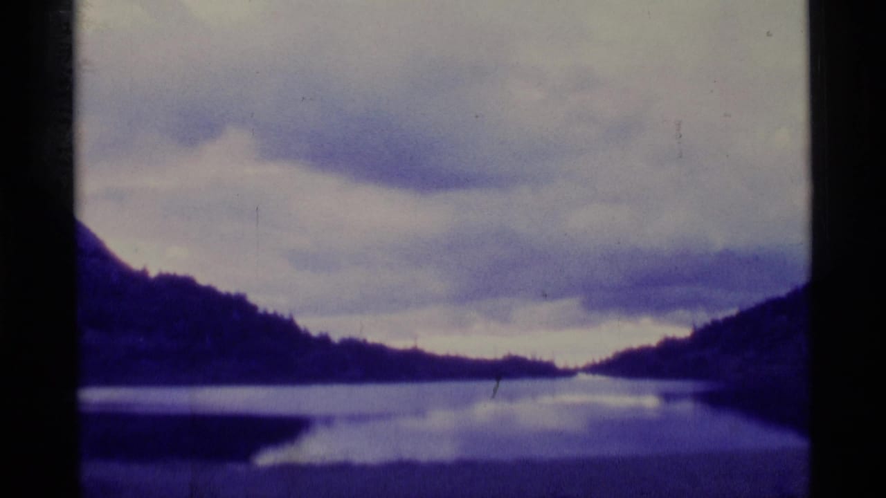 A soft 8mm view of an Alaska lake landscape, with hills reflected in still water under a cloudy sky.