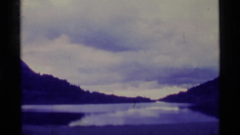 A soft 8mm view of an Alaska lake landscape, with hills reflected in still water under a cloudy sky.