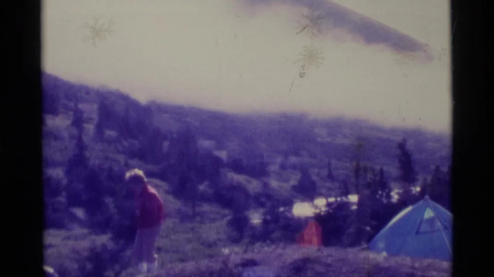 1977 Alaska campsite view with a distant person in a red jacket near a blue tent overlooking lake and hills.