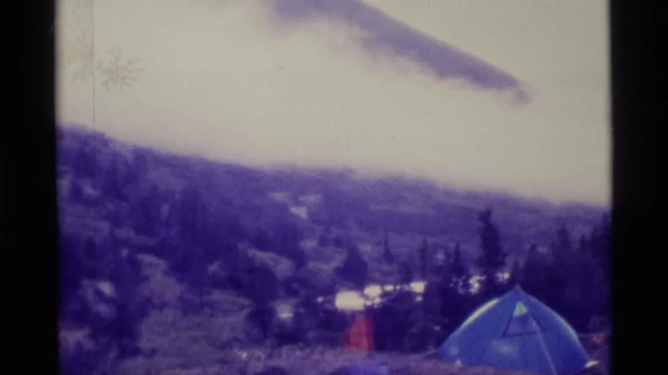A blue tent sits on an Alaska hillside campsite, with water, trees, and clouded hills beyond.