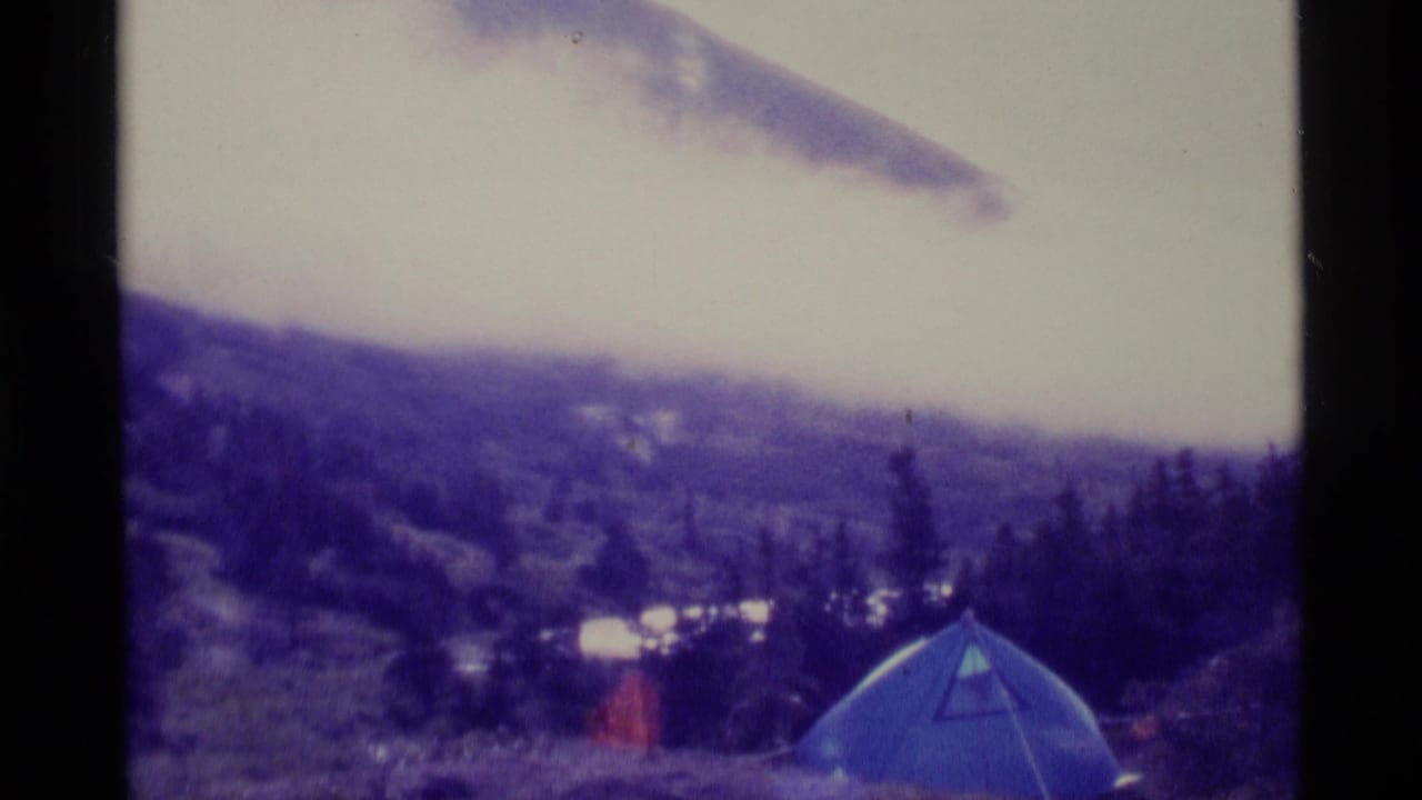 1977 Alaska home-movie frame showing a blue tent pitched on a misty mountain slope with trees and cloudy sky behind it.