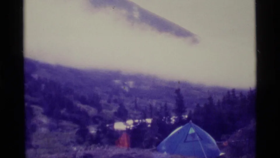 1977 Alaska home-movie frame showing a blue tent pitched on a misty mountain slope with trees and cloudy sky behind it.